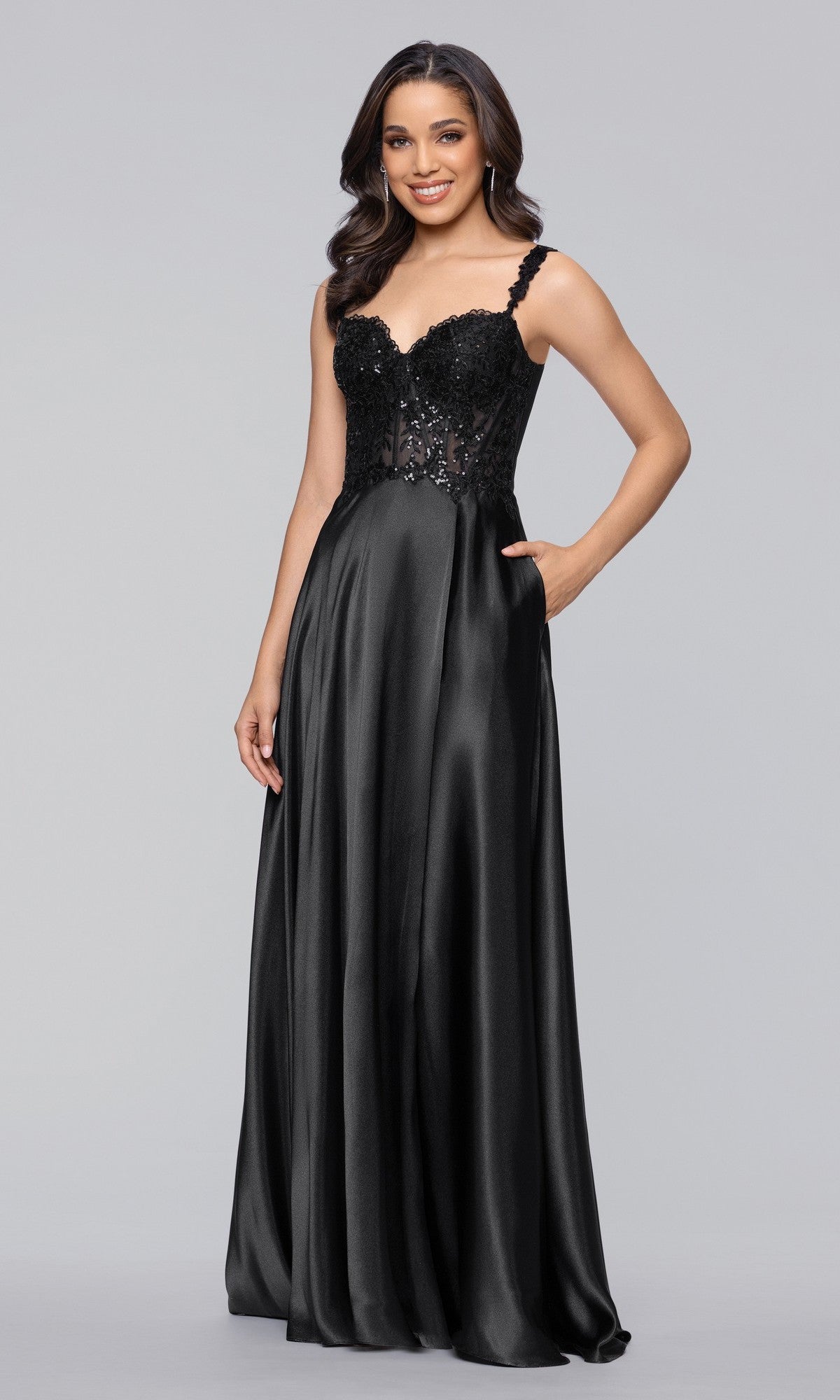 A woman wears the Long Prom Dress: Blondie Nites 6088BN in black, featuring an A-line satin silhouette, sequin-embellished bodice, thin straps, and a flowing skirt, standing against a plain gray background.